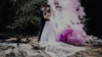 Married couple in tuxedo and dress standing on a rock by a stream with purple smoke behind them
