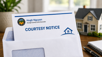 White Envelope with a notice with letter head Hugh Nguyen Clerk Recorder Courtesy Notice and small home. Small model home, white notepad, pen, and green plant in white pot on wooden table with a window and white curtains in the background